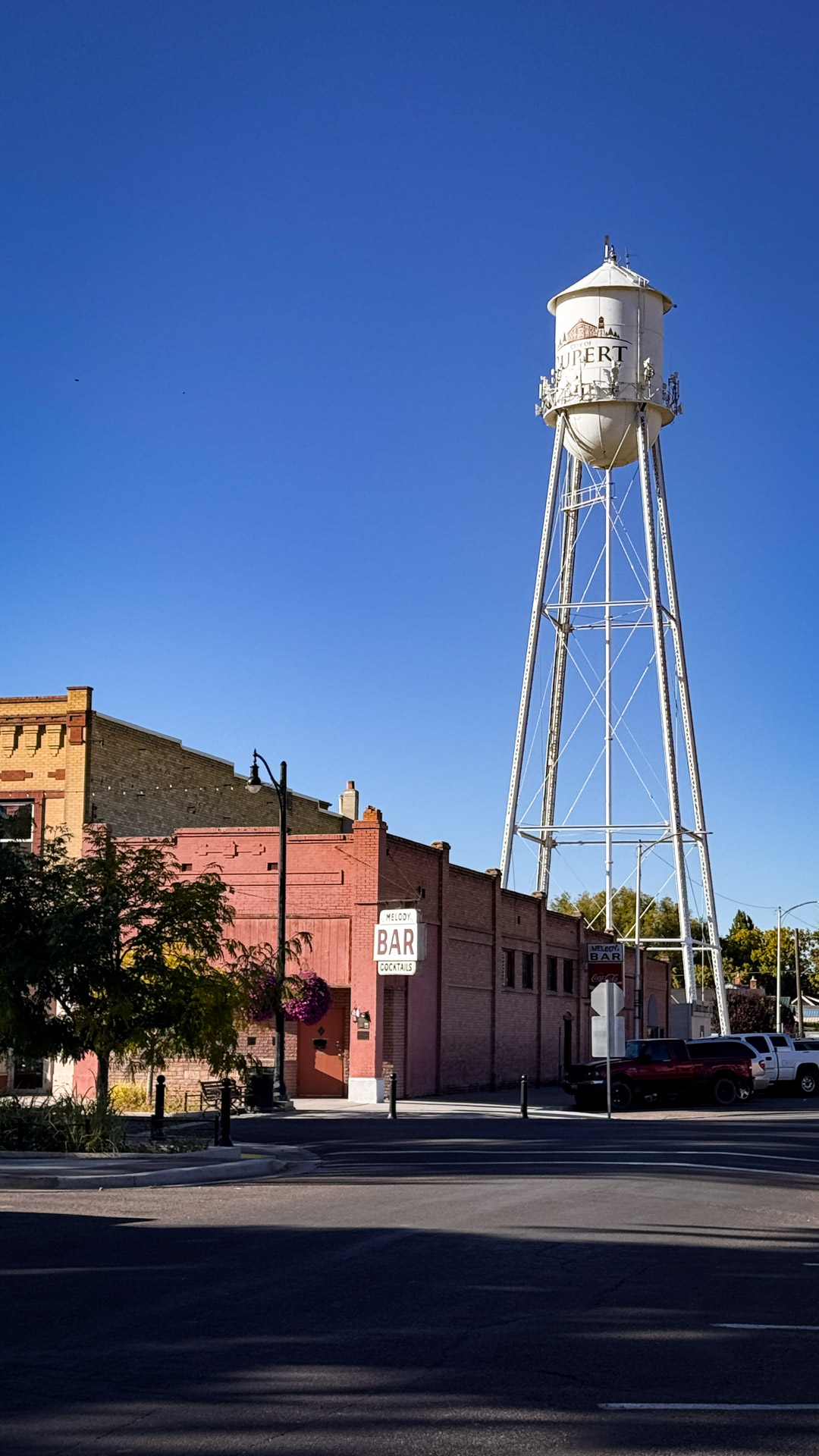 A tall white water tower marked with "ID" stands behind a red brick building with a sign reading Bar Lounge in Rupert. A pickup truck is parked nearby under the clear blue sky.