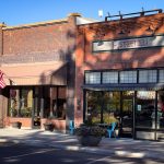 A brick building in Rupert, ID with a sign reading E Street Deli, large windows, blue chairs outside, hanging flower baskets, an American flag, and a sunny sidewalk on a quiet street.