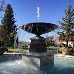 A large, round fountain in Rupert, ID sprays water upwards and cascades into a shallow pool. The fountain is set in a park with trees, flowers, and buildings visible in the background under a clear blue sky.