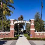 A brick archway labeled Rupert welcomes visitors to the park in Rupert, ID, flanked by flagpoles and trees under a clear blue sky. Circular plaques adorn the brick posts, and a walkway leads into the peaceful grounds.