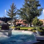 A brick and stone fountain with flowing water sits in sunny Rupert, surrounded by trees, flowers, benches, and shops. A tall white water tower rises in the background against a clear blue Idaho sky.