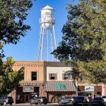 A tall white water tower labeled Rupert, ID, stands behind a row of small brick and wood shops, with trees framing the scene and cars parked in front on a sunny day.