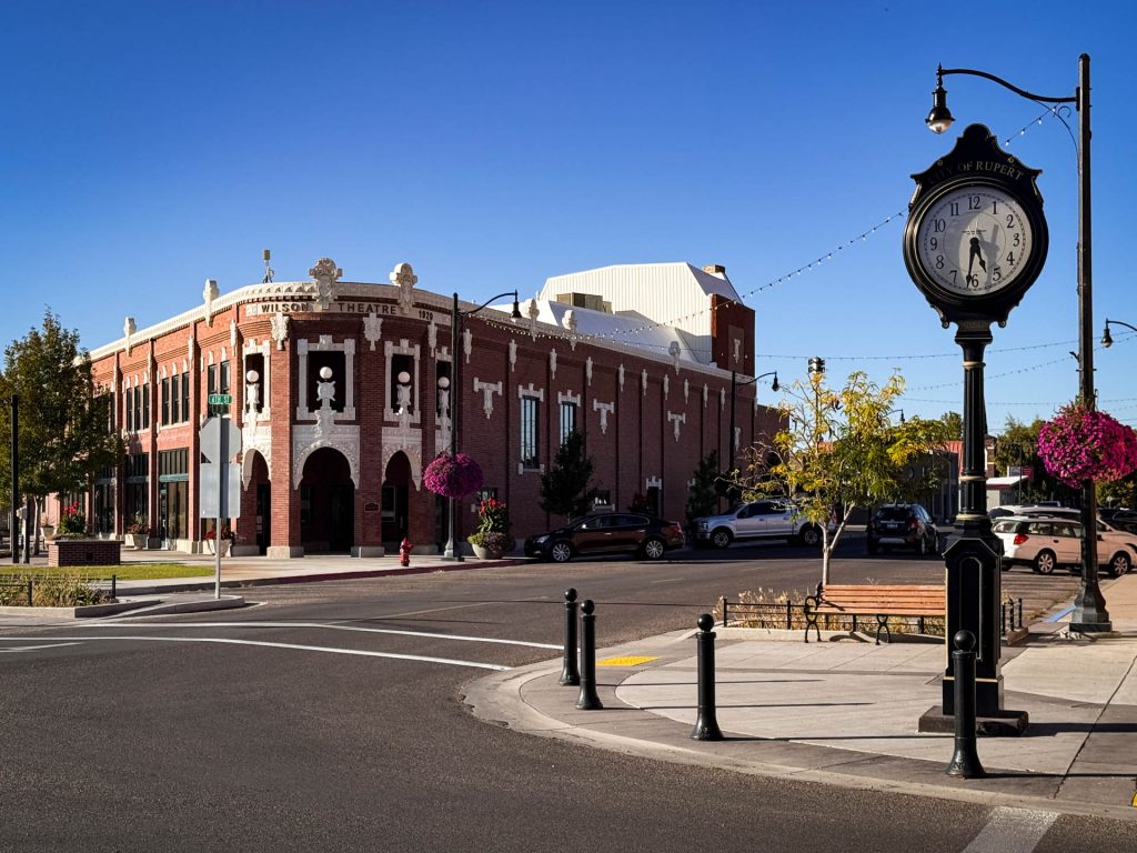 A historic brick building labeled Wilson Theatre sits on a sunny Rupert, ID street corner with parked cars, hanging flower baskets, a vintage clock, and clear blue sky in the background.