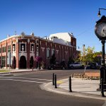 A historic brick building labeled Wilson Theatre sits on a sunny Rupert, ID street corner with parked cars, hanging flower baskets, a vintage clock, and clear blue sky in the background.