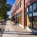 A sunny sidewalk in Rupert, ID runs alongside a row of red brick storefronts with large windows in a small downtown area. Trees, benches, and parked cars line the street, casting shadows on the pavement.