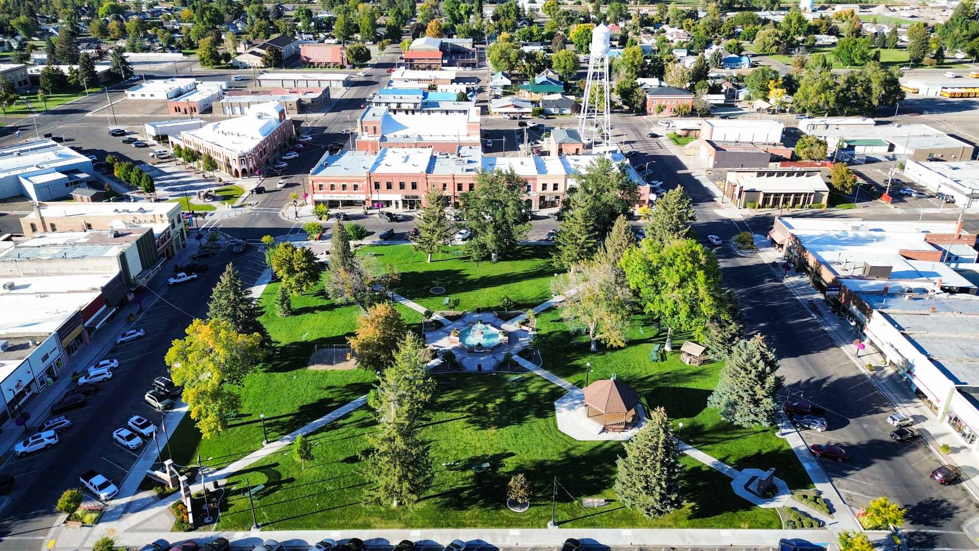 Aerial view of Rupert, ID’s small town park with green grass, trees, walking paths, a gazebo, fountain, and surrounding downtown buildings on a sunny day.