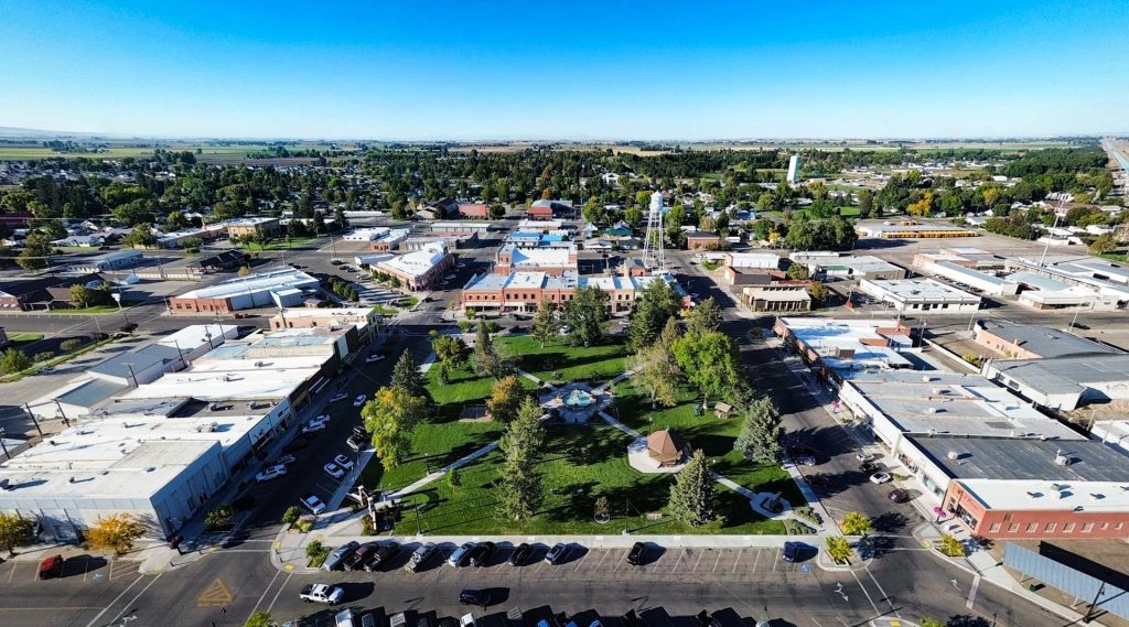Aerial view of Rupert, ID, showing a small town with a central green park, surrounded by trees, buildings, shops, and parked cars under a clear blue sky. Fields stretch into the distance beyond the town.