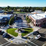 Aerial view of Rupert, ID town square featuring a central monument, green lawns, surrounding sidewalks, parked cars nearby, and historic brick buildings on a sunny day.