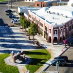 Aerial view of Rupert, ID town square with a brick building, parked cars, green lawn, trees, and a decorative monument in the center. The area features wide sidewalks and street intersections surrounding it.