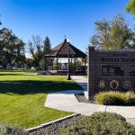 A well-kept park in Rupert, ID, with green grass, trees, a picnic table, and a gazebo. A large brick sign reads “Historic Rupert Square” and lists several community organizations. The sky is clear and blue.