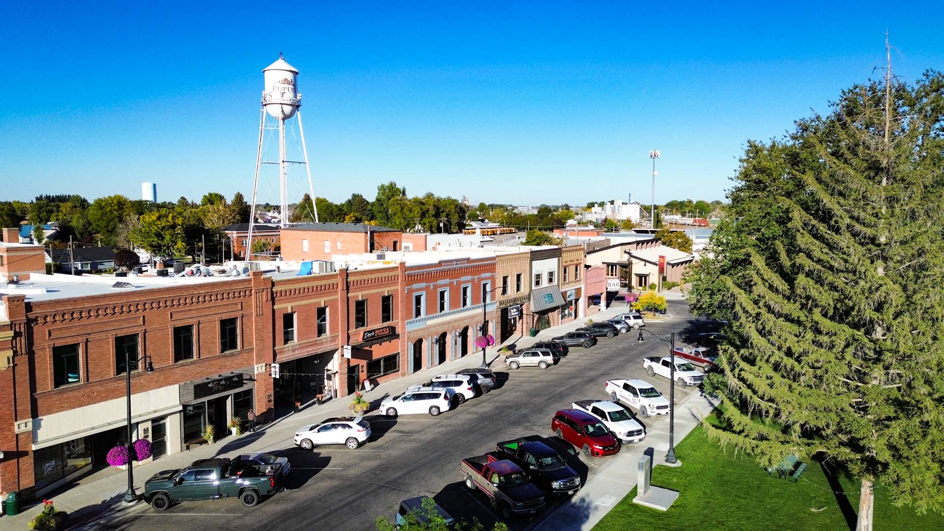 A small town street in Rupert, ID, lined with brick buildings and parked cars. A tall white water tower stands in the background, and trees and a grassy area are visible under a clear blue sky.
