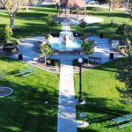 A well-maintained park in Rupert, ID features a central fountain surrounded by benches, pathways, green lawns, trees, and a gazebo in the background on a sunny day.
