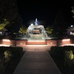 A brightly lit fountain with cascading water stands at the center of a park in Rupert, ID at night, surrounded by trees, benches, and bushes; buildings are visible in the background under a dark sky.