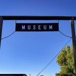 A rectangular black sign with white letters spelling MUSEUM hangs from a wooden frame against a clear blue sky, displaying an ID number attributed to Rupert, with some green trees visible in the lower right corner.