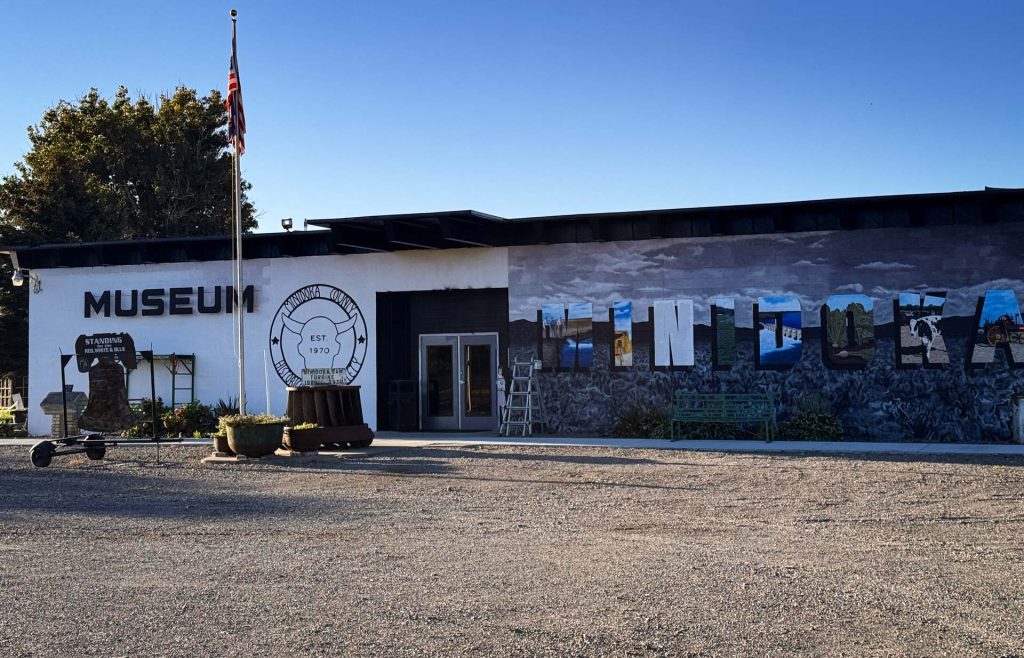 A museum building in Rupert, ID with a mural that says “WINDORA,” featuring blue and gray tones. An American flag flies on a pole outside, surrounded by trees and a clear blue sky in the background.