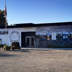A museum building in Rupert, ID with a mural that says “WINDORA,” featuring blue and gray tones. An American flag flies on a pole outside, surrounded by trees and a clear blue sky in the background.