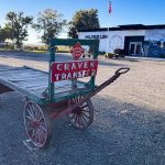 A vintage wooden cart with red wheels and a sign reading CRAVEN TRANSFER sits on gravel in front of a museum building in Rupert, ID, under a blue sky. Trees and a flagpole are visible in the background.