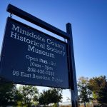A black sign for the Minidoka County Historical Society Museum in Rupert, ID, with hours, phone number, and address, stands against a clear blue sky and trees in the background.
