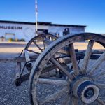 Close-up of a weathered wooden wagon wheel in Rupert, ID, with a museum building blurred in the background under a clear blue sky.