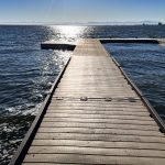 A wooden dock extends into a calm lake under a clear blue sky in Rupert, ID, with sunlight reflecting brightly on the water’s surface and distant mountains visible on the horizon.