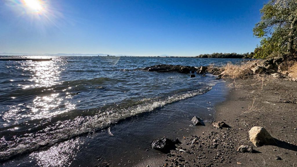 A sunny lakeshore scene near Rupert, ID, with gentle waves lapping against a rocky, sandy beach. The water sparkles in the sunlight, and trees line the shore beneath a clear blue sky.
