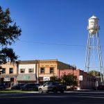 A small town street in Rupert, ID features historic brick buildings, parked cars, and a tall white water tower labeled Rupert under a clear blue sky. Trees and string lights add charm to this inviting scene.